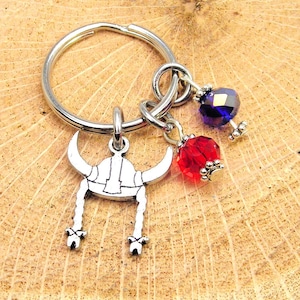 May include: A silver-toned keychain featuring a Viking helmet charm with dangling braids, paired with red and blue faceted bead accents. The keychain is attached to a silver ring, perfect for keys or bags.