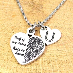 Op de afbeelding: Zilveren ketting met twee hartvormige hangers. Een hart heeft de tekst "De helft van mijn hart leeft in de hemel" en een engelenvleugel. Het andere hart heeft de letter "U" gegraveerd.