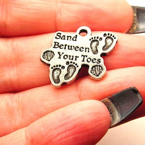 May include: A silver-colored charm with the words "Sand Between Your Toes" and footprints and seashell designs. The charm has a small loop at the top for attaching to a necklace or bracelet. The charm is held in a hand.