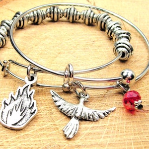 May include: Two silver bangle bracelets with charms. One charm is a silver flame, another is a silver dove, and the third is a red crystal.