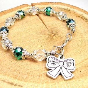 Puede incluir: Un brazalete de plata con cuentas de vidrio verde y transparente. El dije es un lazo de plata con el texto "Working My Bow Off".