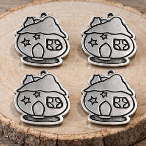 May include: Four silver-colored house charms with star and window details, arranged on a wooden slice. Each charm has a loop at the top for attaching to jewelry or other crafts. The charms are detailed with black outlines.