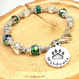 Op de afbeelding: Een groene en heldere kristallen armband met een zilveren bedel met de tekst "Wildcats" en een pootprint.