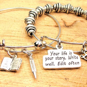 Puede incluir: Dos pulseras plateadas con dijes. Una tiene un cristal rojo y un dije de libro. La otra tiene cuentas plateadas y un dije que dice "Your life is your story. Write well. Edit often." y un dije de "Love".