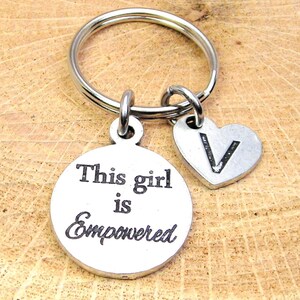 Könnte beinhalten: Silberner Schlüsselanhänger mit einem runden Anhänger, auf dem "This girl is empowered" steht, und einem Herzanhänger mit dem eingravierten Buchstaben "V".