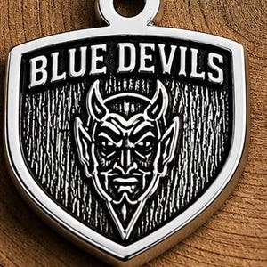 May include: A silver-colored pendant with the words "BLUE DEVILS" at the top. The pendant features a devil face in the center. The pendant is shield-shaped with a loop at the top for a chain.