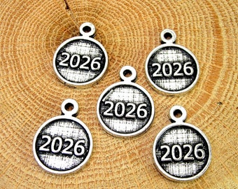 Könnte beinhalten: Fünf silberne Anhänger mit dem Jahr 2026, das auf jedem Anhänger geprägt ist. Die Anhänger sind rund und haben eine strukturierte Oberfläche.
