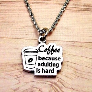 May include: Silver necklace with a pendant displaying a coffee cup illustration and the words "Coffee because adulting is hard." The pendant hangs from a silver rope chain, ideal for coffee enthusiasts.