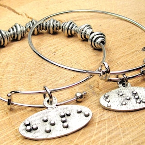 May include: Two silver charm bracelets with a beaded charm on one and a silver charm with the word "LOVE" written in braille on the other. The second bracelet has a red crystal charm.