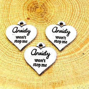 Könnte beinhalten: Drei silberne herzförmige Anhänger mit dem Text "Anxiety won't stop me" auf jedem Anhänger. Die Anhänger liegen auf einer Holzoberfläche.