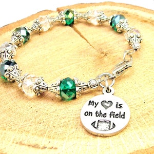 Puede incluir: Pulsera de cuentas con cuentas facetadas transparentes y verde esmeralda, intercaladas con adornos decorativos de metal plateado. Un dije plateado con el texto "My heart is on the field" y un gráfico de fútbol americano cuelga.