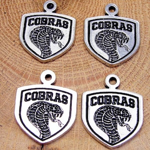 Könnte beinhalten: Vier silberfarbene Metallanhänger in Schildform. Jeder Anhänger zeigt das Wort "COBRAS" über einer detaillierten Illustration einer Kobra-Schlange. Die Anhänger haben oben eine Schlaufe zum Befestigen.