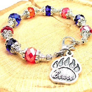 Puede incluir: Un brazalete de plata con cuentas de cristal facetado rojo, azul y naranja. El brazalete tiene un cierre de plata y un dije de plata con una huella de pata de oso y la palabra "Bears" inscrita en él.