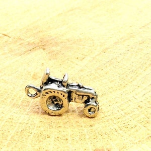 May include: A silver charm in the shape of a tractor. The tractor has a single large wheel and a smaller wheel on the back. The charm is detailed and has a realistic design.