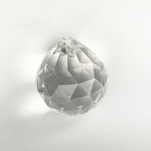 Op de afbeelding: Een heldere, gefacetteerde kristallen bol met een lus aan de bovenkant om op te hangen. Het kristal is gesneden met talrijke geometrische vormen, die licht reflecteren. Het object is decoratief.