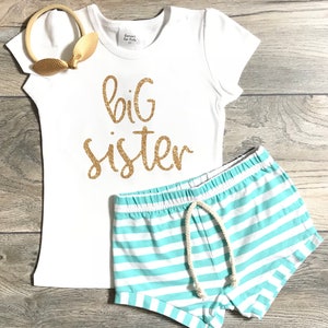 May include: White t-shirt with gold glitter text that says "big sister" and matching light blue and white striped shorts.