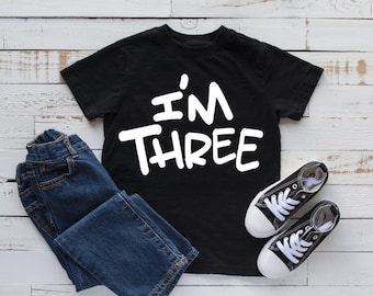 Może przedstawiać: Czarny t-shirt z białym napisem "I'm Three". Koszulka leży na białym drewnianym tle z niebieskimi dżinsami i białymi butami sportowymi.