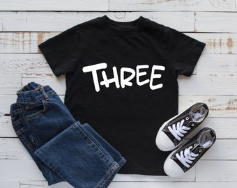 Może przedstawiać: Czarny t-shirt z białym napisem "THREE". Koszulka leży na białym drewnianym tle z niebieskimi dżinsami i czarnymi butami sportowymi.