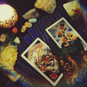 May include: A tarot card reading spread with a variety of cards, including the Page of Earth, the XII card, and the 21 card. The cards are laid out on a dark surface with crystals and a candle.