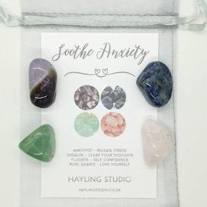 May include: A set of five tumbled stones in a white organza bag. The stones are amethyst, sodalite, fluorite, rose quartz, and a green stone. The bag has a white card with the text "Soothe Anxiety" and a list of the stones and their properties.
