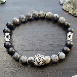 May include: A beaded bracelet featuring a mix of black and gray stone beads. The bracelet has silver-colored metal accents, including two cylindrical pieces and a central skull-shaped bead. The bracelet is displayed on a dark wooden surface.