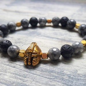 May include: A gray and black beaded bracelet with a gold gladiator helmet charm. The bracelet is made with lava rock and gray gemstone beads.