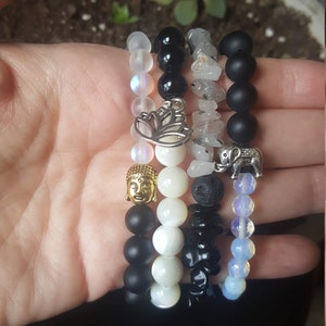 Puede incluir: Un conjunto de cinco pulseras con diferentes cuentas y dijes. Las pulseras incluyen cuentas negras, blancas y transparentes, así como un dije de Buda, un dije de flor de loto y un dije de elefante.