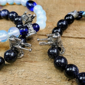 Puede incluir: Dos pulseras con dijes de plata con forma de Daleks, un robot ficticio de la serie de televisión de ciencia ficción británica Doctor Who. Una pulsera está hecha de cuentas azules y blancas, la otra está hecha de cuentas negras con motas plateadas.