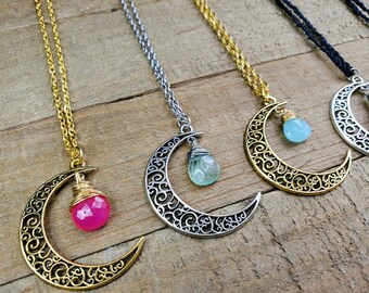 May include: Four necklaces with crescent moon pendants and different colored gemstone drops. The necklaces are on a wooden surface. The gemstones are pink, blue, and green.