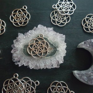 May include: A collection of silver metal charms featuring a flower of life design, displayed on a purple and white amethyst crystal and a crescent moon shaped amethyst crystal.