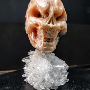Peut inclure: Une sculpture de crâne sculpté en tons pêche et beige, montée sur un amas de cristaux de quartz clairs. Le crâne présente des détails, notamment des orbites, un nez et une bouche. Les cristaux sont disposés en forme irrégulière et épineuse.