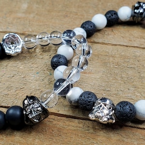 May include: Two beaded bracelets with a Star Wars theme. One bracelet features black matte onyx, clear quartz, white howlite, and lava rock beads. The other bracelet has similar beads. Both bracelets have silver-toned character charms.