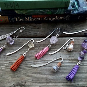 May include: Six silver bookmark pendants with different charms and tassels. The charms include a feather, a clear crystal, a heart-shaped crystal, a small bottle with a cork, and a strand of amethyst stones. The tassels are in shades of purple, orange, and pink.