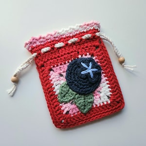 May include: A handmade red crochet drawstring bag with a blueberry design. The bag features a dark blue blueberry with a light blue cross-shaped center, green leaves, and a pink and white floral pattern. The drawstring is white with wooden beads.