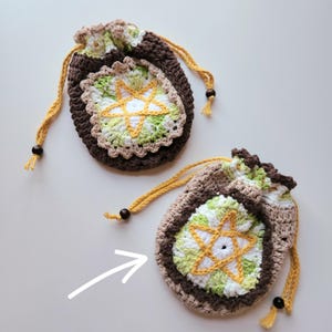 May include: Two handmade crochet drawstring pouches. The pouches are brown, beige, and green, with a starburst design in the center. The drawstring is yellow with a wooden bead. The pouches are likely used for small items or gifts.