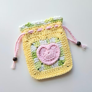 May include: A handmade crochet drawstring bag in a square shape. The bag features a yellow base with a pink heart applique in the center. The bag has a pink drawstring with wooden beads and a scalloped edge.