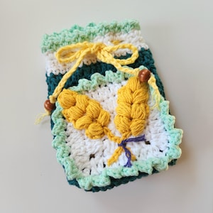 May include: A handmade crochet pouch in shades of green, white, and teal, with a yellow drawstring and two yellow wheat-like embellishments. The pouch features a square base and a scalloped edge, with a small purple bow.
