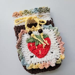 May include: A handmade crochet pouch featuring a red strawberry applique. The pouch is white with a colorful scalloped edge and a brown base. It has a yellow drawstring closure and two brown bead accents.