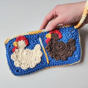 May include: A handmade crochet pouch featuring two chickens. One chicken is white, the other is brown, both wearing red Santa hats. The pouch is blue with a yellow and cream border and a cream and yellow strap.