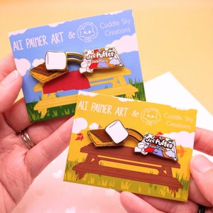 May include: Two enamel pins featuring a s'more with a Jet-Puffed marshmallow and a chocolate bar on a picnic table. The pins are on cards with the text "Ali Palmer Art & Cuddle Sky Creations".