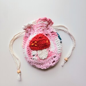 May include: A pink and white crocheted drawstring bag with a red mushroom design. The bag has a white cord with wooden beads.