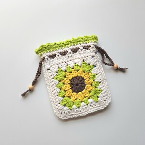 May include: A crocheted drawstring bag featuring a sunflower design. The bag is primarily off-white with a large sunflower in the center, using yellow, green, and brown yarn. The top edge is green, and the drawstring is brown with wooden beads.