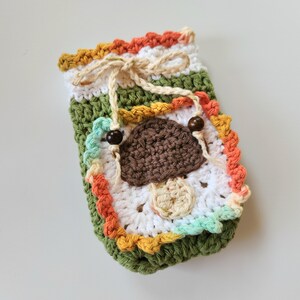 May include: A handmade crochet pouch in green, white, and orange, featuring a brown mushroom applique. The pouch has a drawstring closure with wooden beads and a decorative border. The overall design is whimsical and nature-inspired.