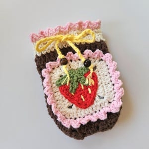 May include: Crocheted strawberry-themed pouch. The pouch features a red strawberry applique with green leaves on a cream-colored square. The pouch has a brown base, pink and cream trim, and a yellow drawstring with wooden beads.