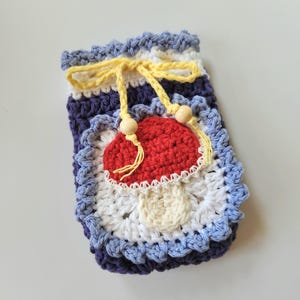 May include: A handmade crochet pouch in shades of blue, white, and purple, featuring a red mushroom applique. The pouch has a yellow drawstring closure with wooden beads. The design is a whimsical, handcrafted item.