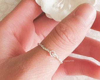May include: A silver chain ring worn on a finger. The ring is delicate and has a simple design. The ring is worn on a hand holding a white paper flower.