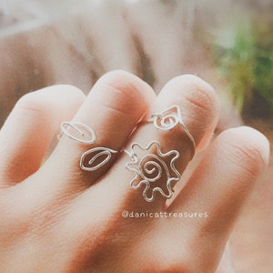 May include: A set of three silver wire rings featuring a sun, a leaf, and a swirl design. The rings are worn on a person's fingers.