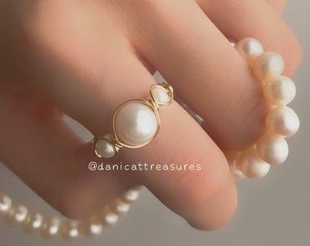 May include: A gold ring with a pearl design. The ring is wrapped with wire and features three pearls. The ring is being held by a hand wearing a pearl necklace.