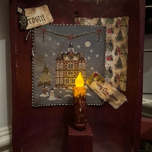 Puede incluir: Una cuchara de madera rústica de color burdeos exhibe una escena de punto de cruz de un pueblo invernal con una vela encendida. El punto de cruz presenta una casa, un muñeco de nieve y un árbol de Navidad. Las etiquetas dicen "Frosty" y "Merry Christmas".