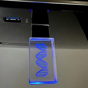Könnte beinhalten: Ein klarer, rechteckiger Block mit einem blau-gelben DNA-Strang-Design. Der Block wird mit blauem Licht beleuchtet, wodurch ein Leuchteffekt entsteht. Das Objekt hängt vor einem dunklen Hintergrund.